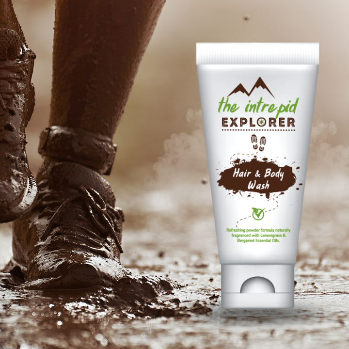 A tube of powdered hair and body wash by The Intrepid Explorer placed beside a person's feet stepping into mud, emphasizing its use for outdoor activities.
