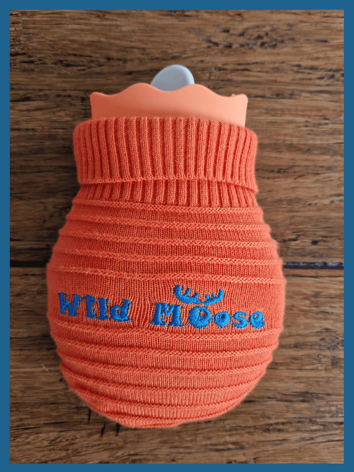 brightly coloured mini hot water bottle in a knitted cover in orange against a wooden floor backdrop