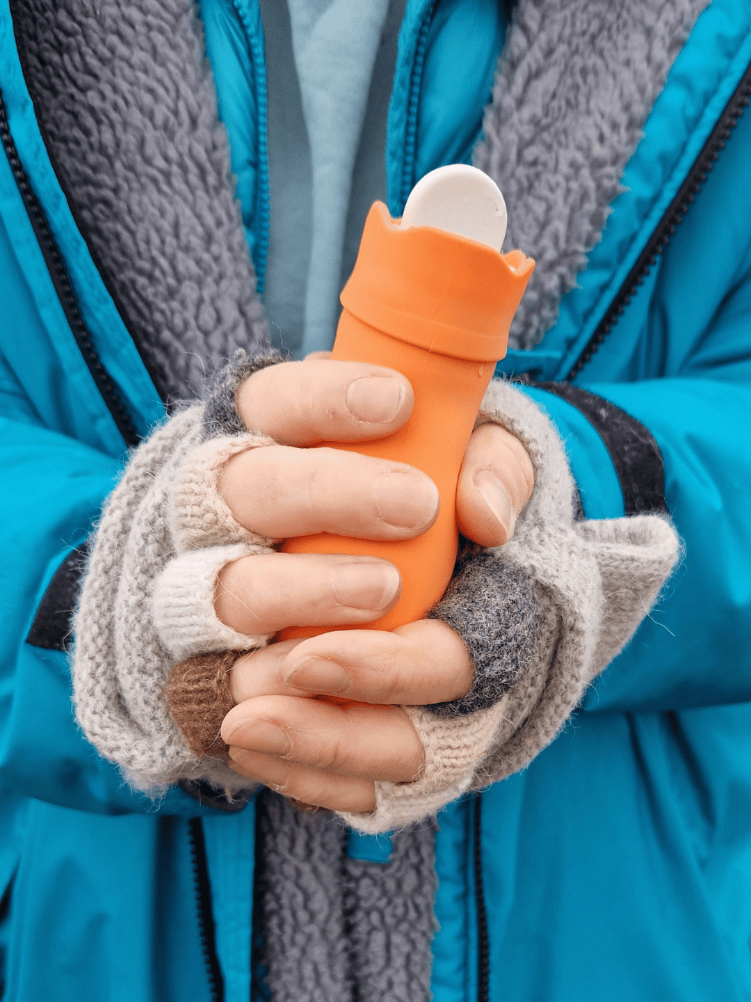 close up of a brightly coloured mini hot water bottles in orange held by hands wearing grey fingerless gloves