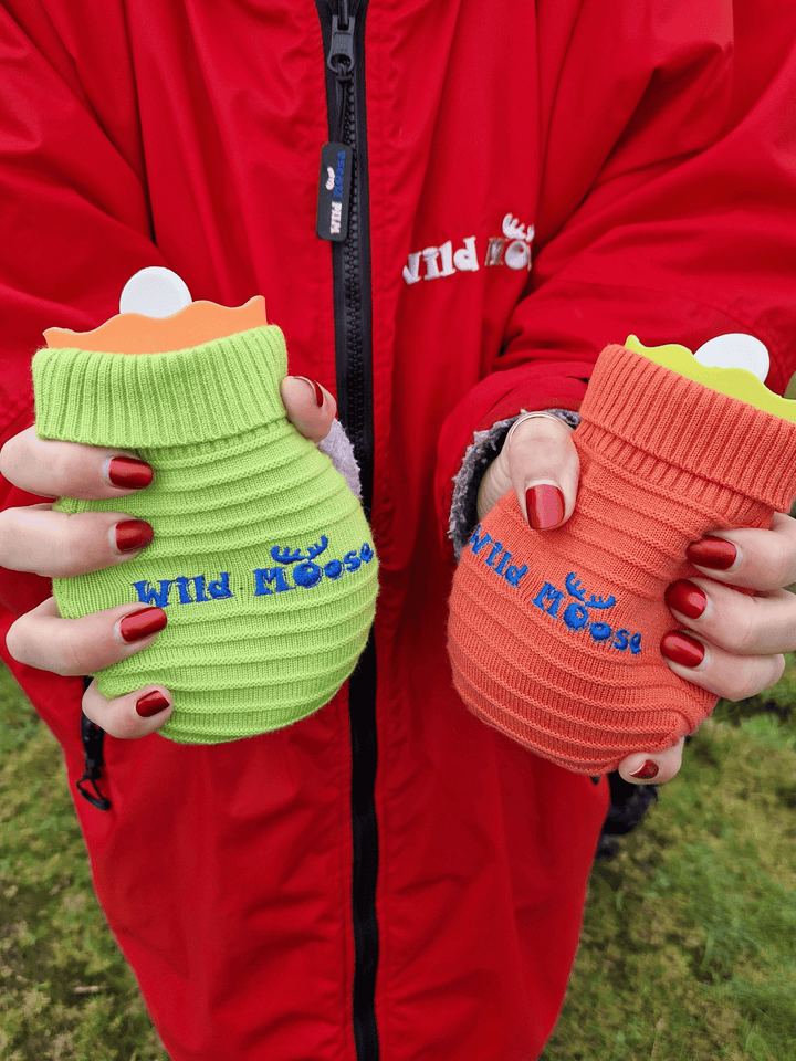 brightly coloured mini hot water bottles in a knitted cover in orange and lime green - held by a lady wearing a red changing robe and with red nail polish on her fingernails