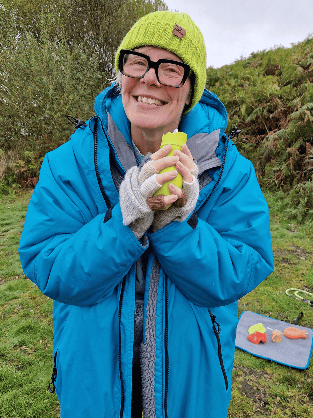 brightly coloured mini hot water bottle in a knitted cover in lime green held by a smiling lady wearing a woollen hat of the same colour and a teal coloured changing robe 