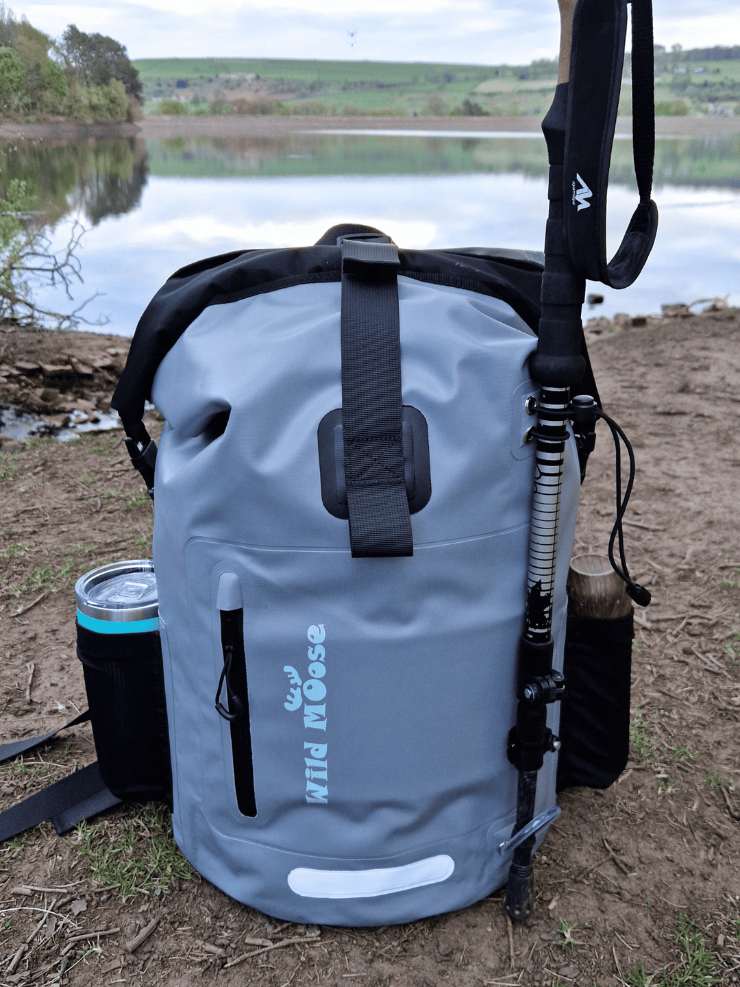 a light grey backpack on the ground in front of a reservoir. 2 flasks in the side pockets and a single walking pole on the front