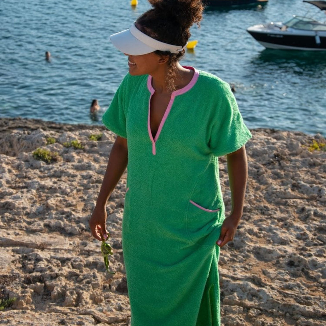 A woman standing on a beach wearing a green knee-length cover-up dress with pink trim, in soft cotton towelling material