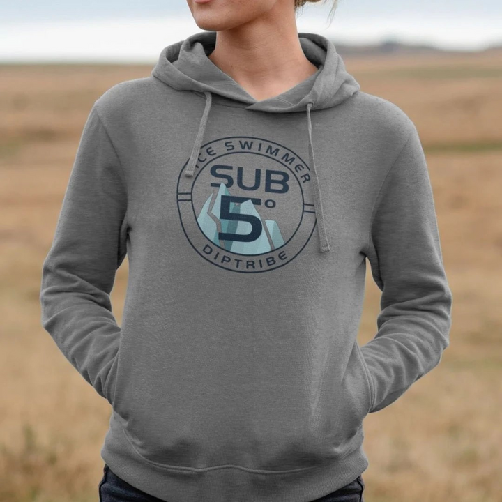 A woman standing in a field wearing a grey diptribe hoodie with an 'Ice swimmer SUB 5' circular logo on the front.