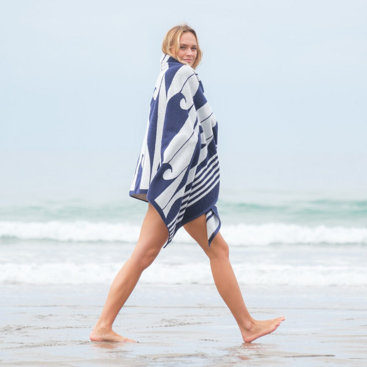 Woman walking on a beach with a blue and white patterned towel draped over her shoulders.