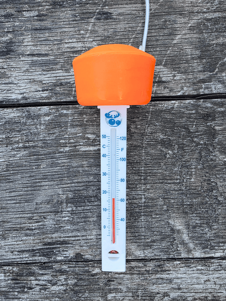 A floating thermometer with a bright orange top and a white plastic body, showing temperature scales in Celsius and Fahrenheit. The thermometer is photographed against a weathered grey wooden plank background.