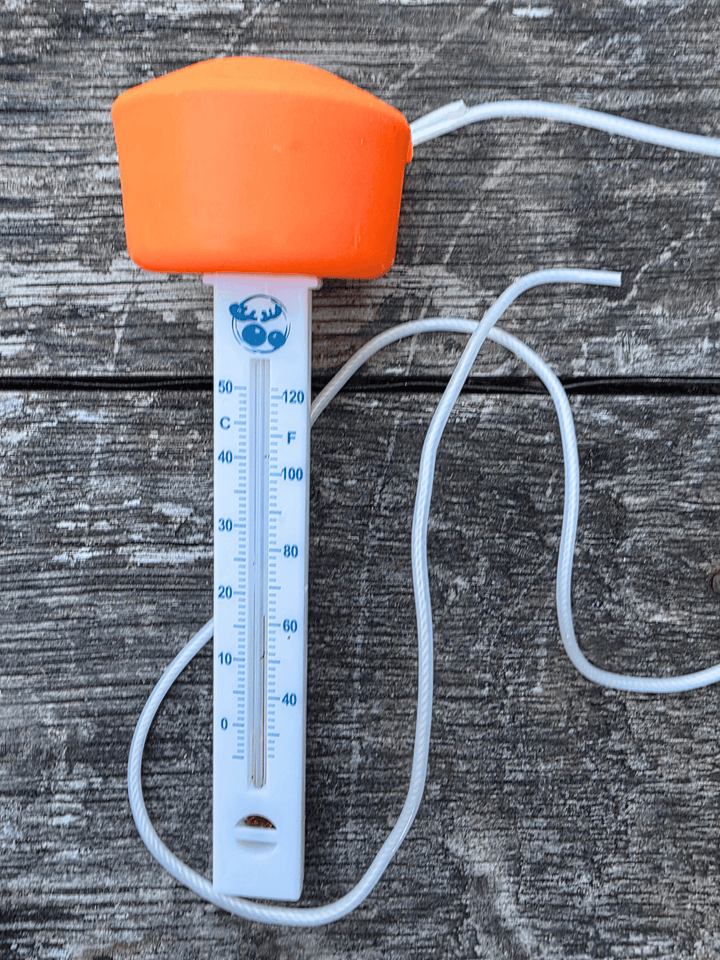 An outdoor swimming thermometer with a bright orange cap, white plastic body, and white cord, shown on a weathered grey wooden surface.