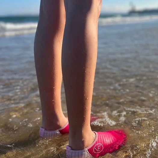 Lifestyle image of a woman standing in the sea wearing a pair of the VE Shoe hot pink water shoes with white sole.