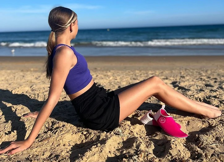 Lifestyle image of a young woman sitting on a beach with her toes in the sand and a pair of VE hot pink water shoes in the sand beside her