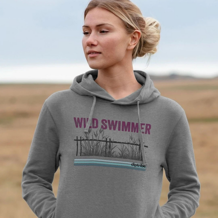 A lifestyle image of a woman standing in a field wearing a diptribe slim-fit hoodie in light heather with the text 'Wild Swimmer' and a hand-drawn graphic of reeds on the front.