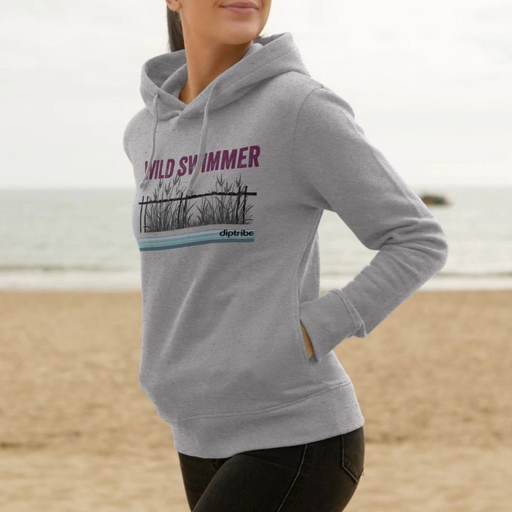 A lifestyle image of a woman on a beach wearing a diptribe slim-fit hoodie in light heather with the text 'Wild Swimmer' and a hand-drawn graphic of reeds on the front.
