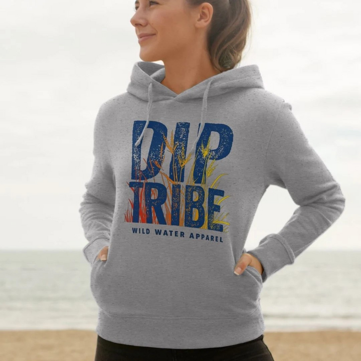 A lifestyle image of a woman standing on a beach wearing a diptribe slim-fit hoodie in light heather with the text 'Diptribe Wild Water Apparel' and a hand-drawn colourful graphic of reeds on the front.
