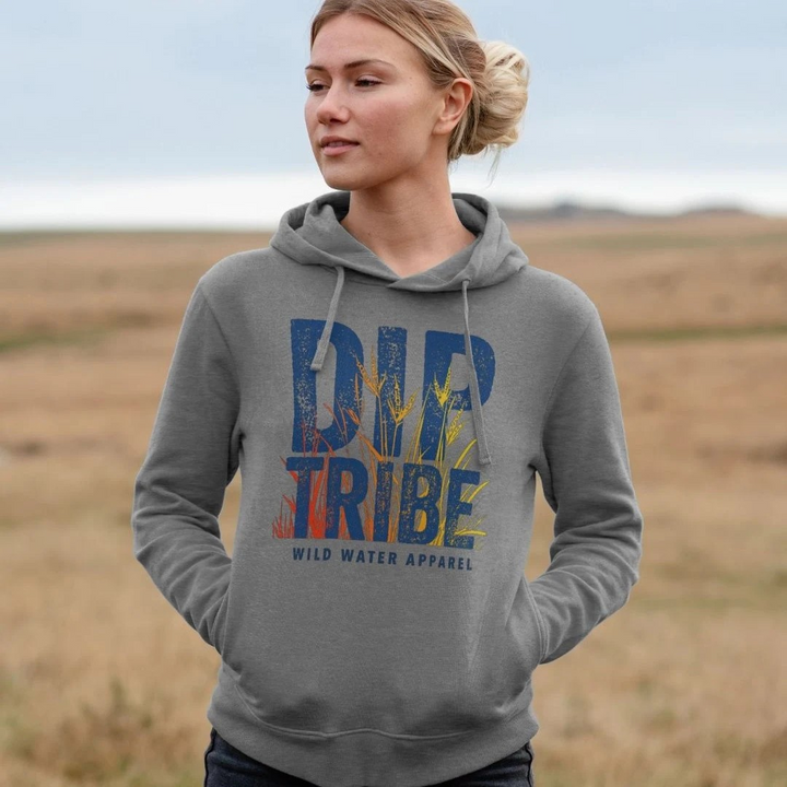A lifestyle image of a woman standing in a field wearing a diptribe slim-fit hoodie in light heather with the text 'Diptribe Wild Water Apparel' and a hand-drawn colourful graphic of reeds on the front.