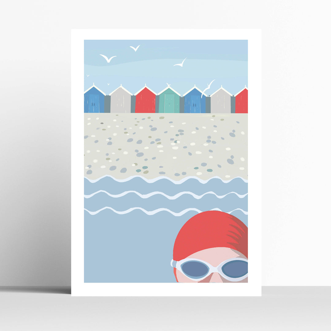 A4 art print featuring a beach scene with colourful beach huts, pebbled sand, gentle waves, and a swimmer in a red swim cap and goggles at the bottom right, shown against a light gray background.