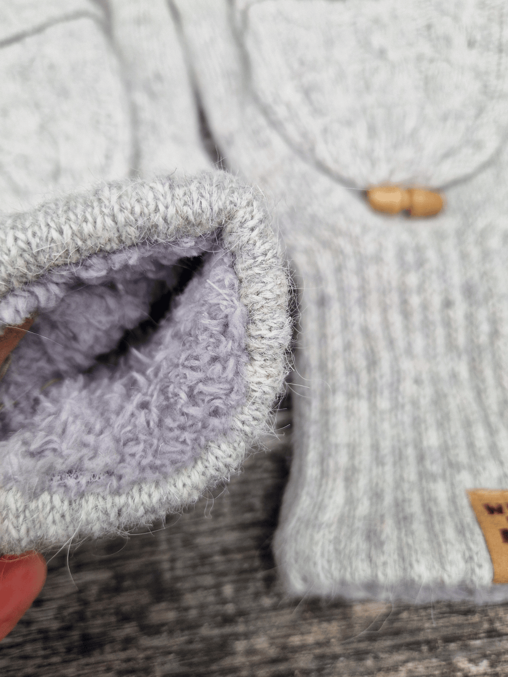 Pair of grey fingerless gloves with wooden buttons on a wooden surface. There is a folded back mitten cover. Close up of the fleece lining at the wrist cuff