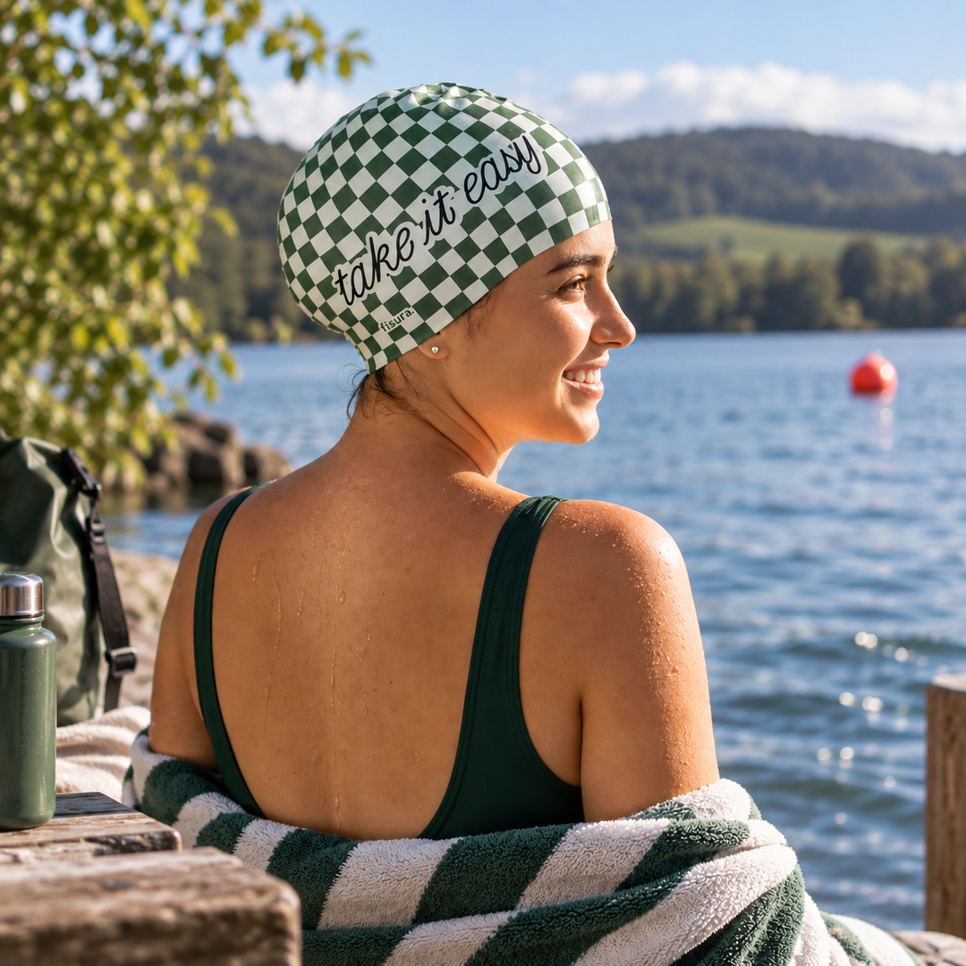 A woman sitting by a lake wearing a green and white checkered swim cap with the words &