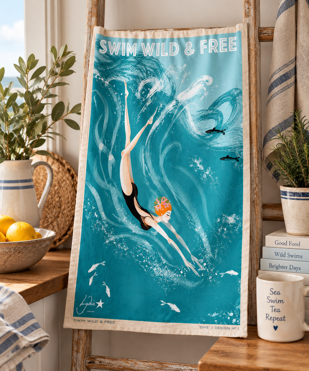 A vintage-style illustrated towel hanging on a wooden ladder in a bright kitchen setting. The towel features a teal and turquoise ocean scene with a diver, fish, and the text 'SWIM WILD & FREE' at the top.