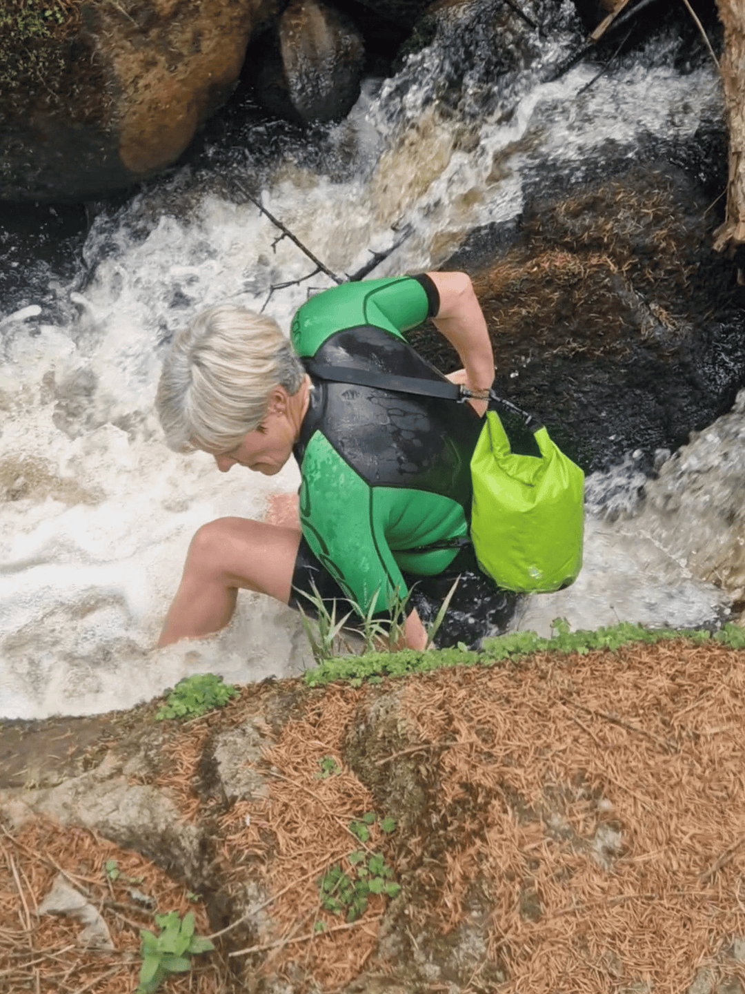 lady in wetsuit in waterfall with 5L green dry bag over shoulder. View from above
