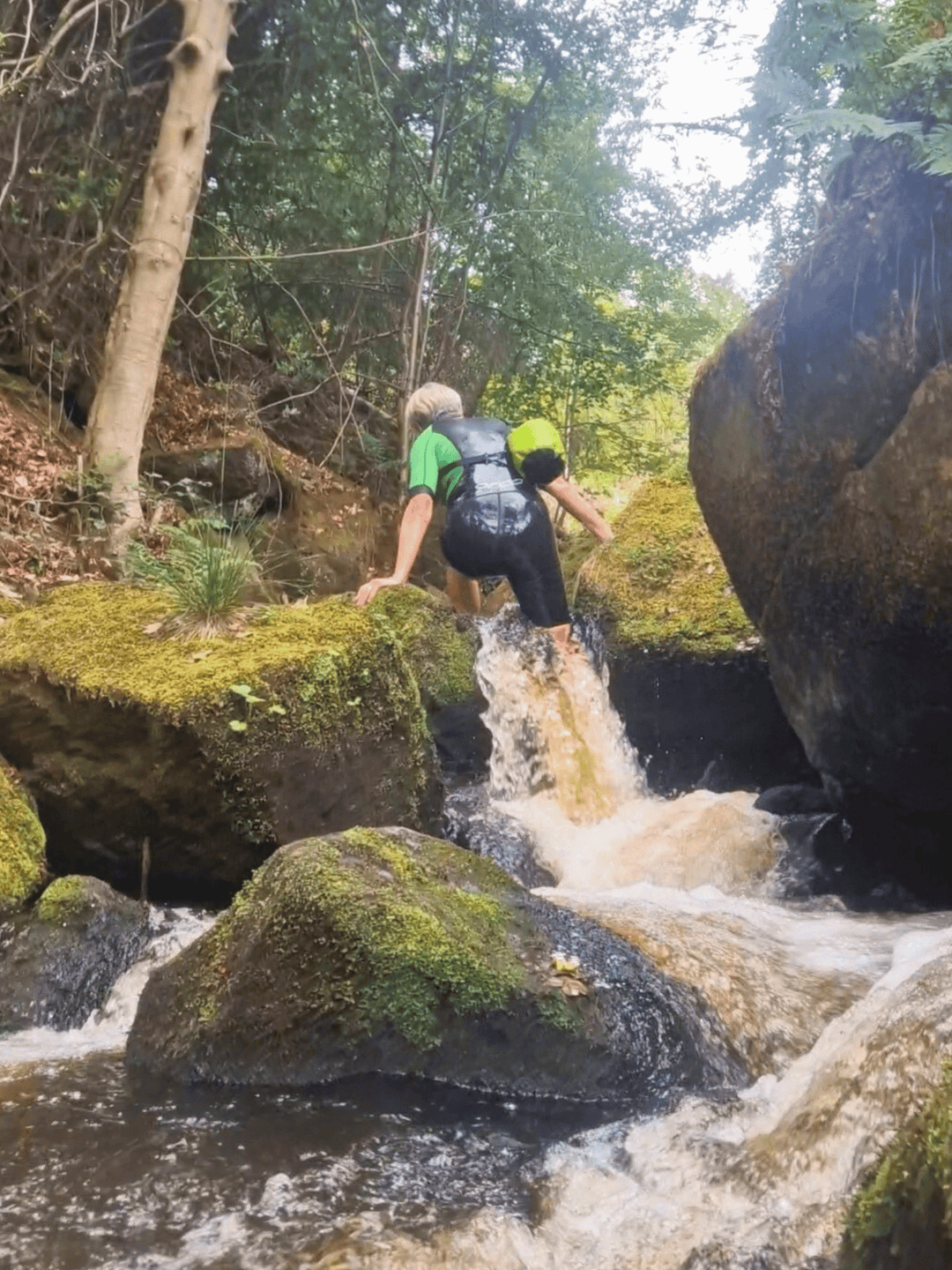 lady in wetsuit stepping out of small waterfall. She has green 5L dry bag over shoulder