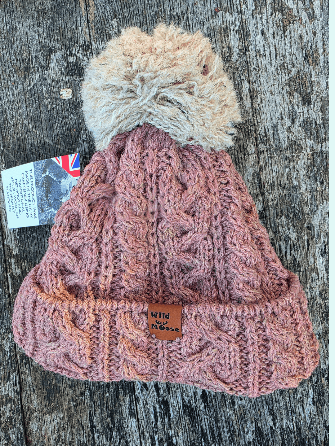 Dusty pink knitted bobble hat with a beige pom-pom on a wooden surface