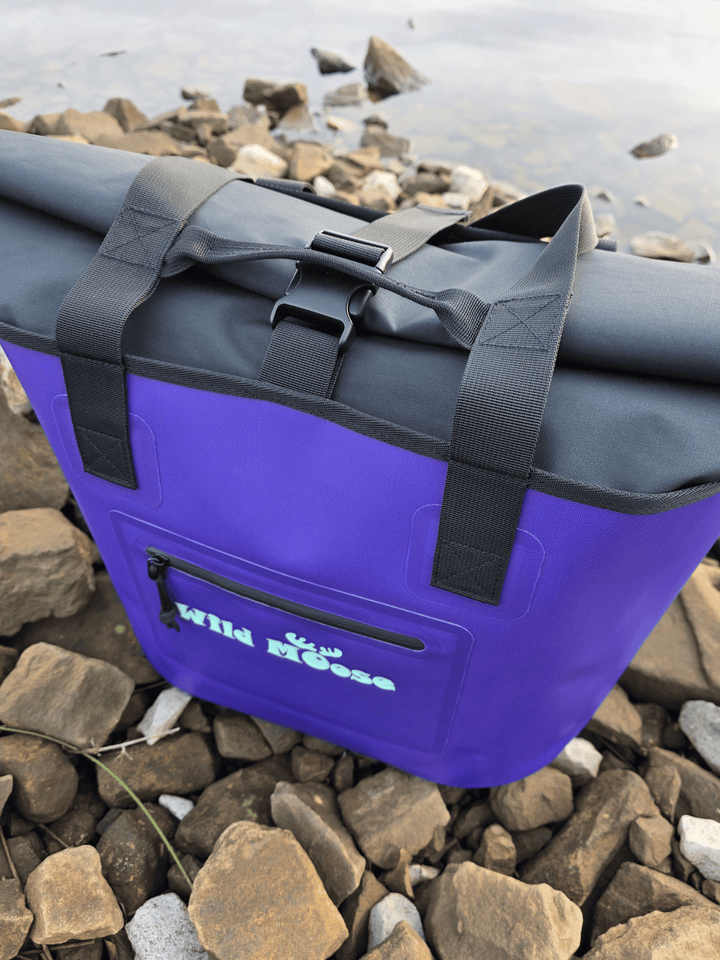 Purple tote bag with black roll top on rocks near water