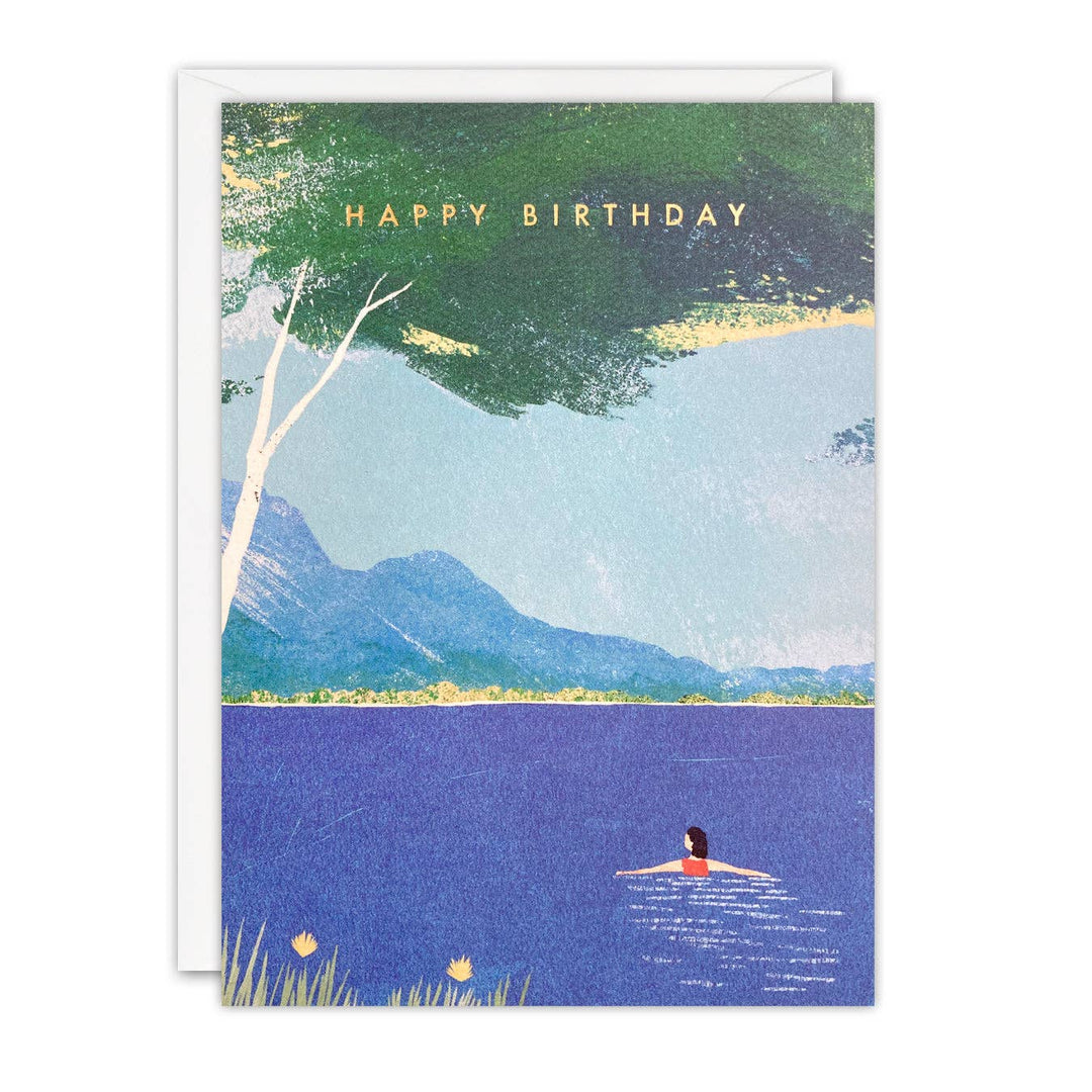 Lake Swimming birthday card illustrated by Henry Rivers featuring a lone swimmer in a blue lake beneath a tree with mountains in the background and Happy Birthday in gold foil.