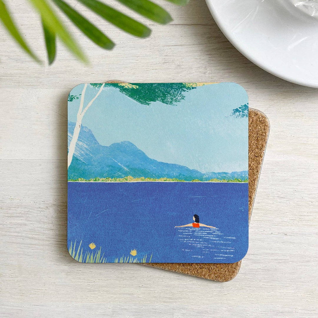 Lake Swimming cork-backed coaster with an illustration by Henry Rivers displayed on a table in a home setting.