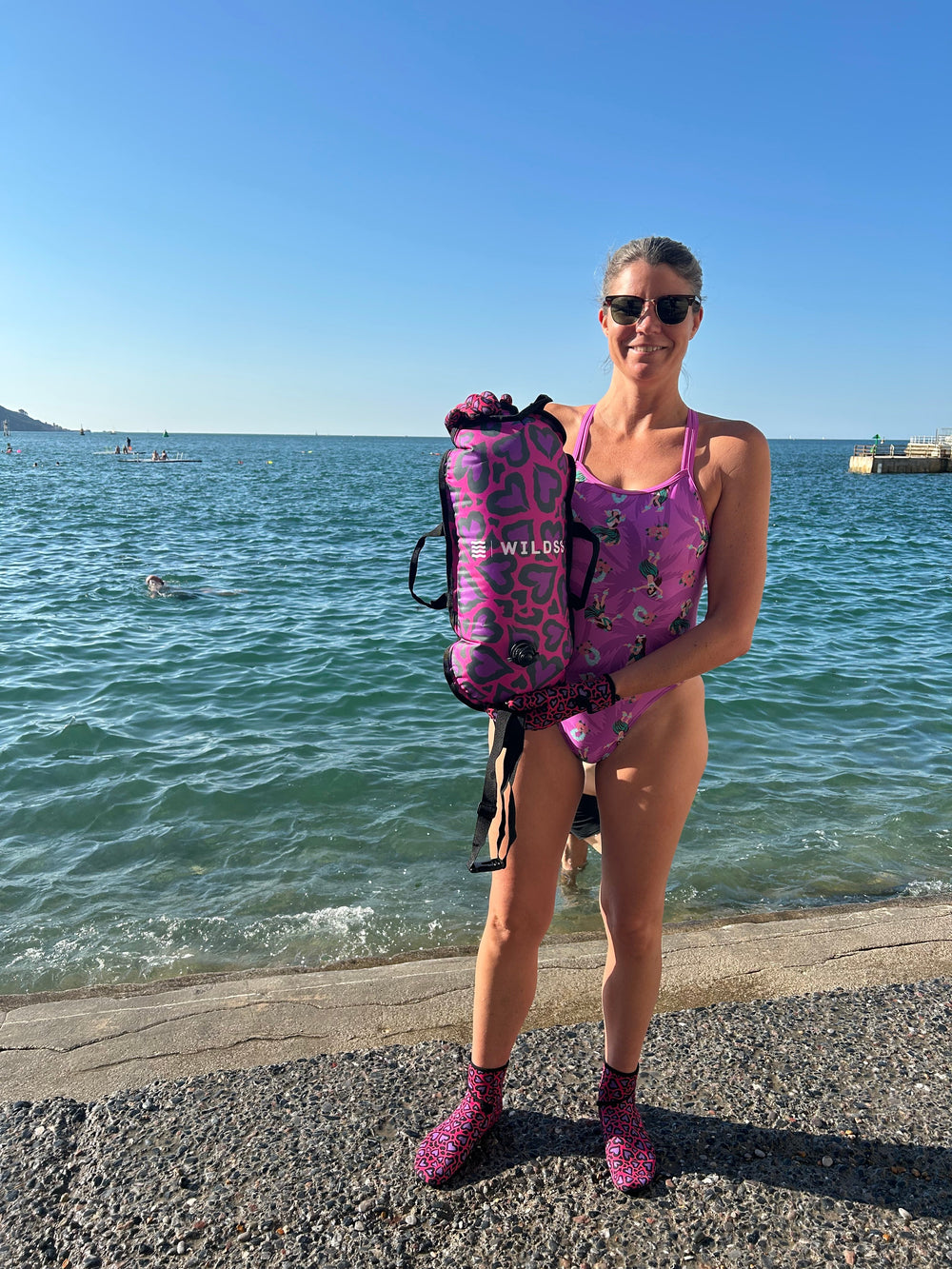 Open water swimmer standing at water's edge holding a pink Wild Swimming Socks Cold Cold Heart print swim buoy with built-in thermometer.
