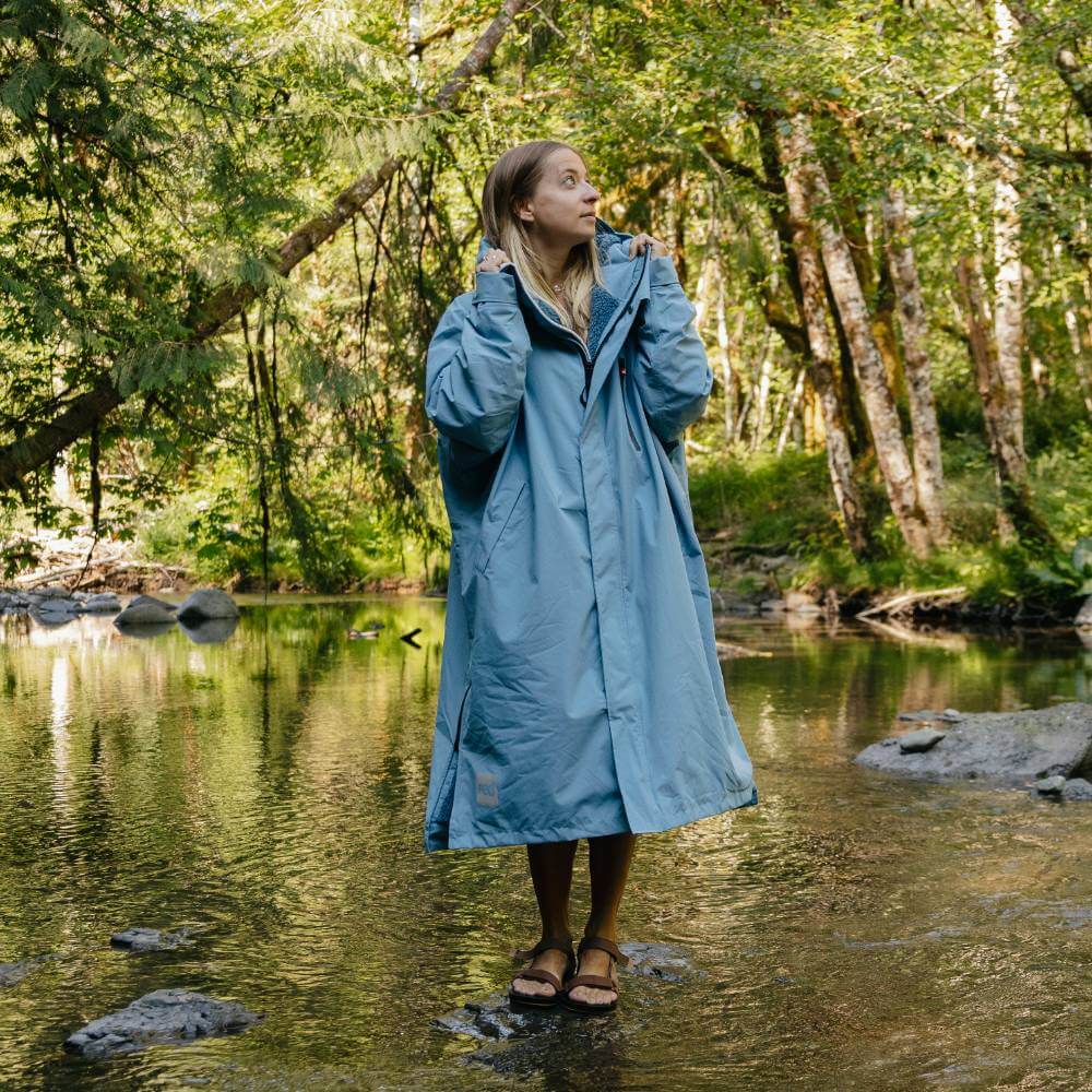 Woman standing on a stepping stone in a river wearing a Red Alter Evo long sleeve waterproof changing robe in Alpine Blue.