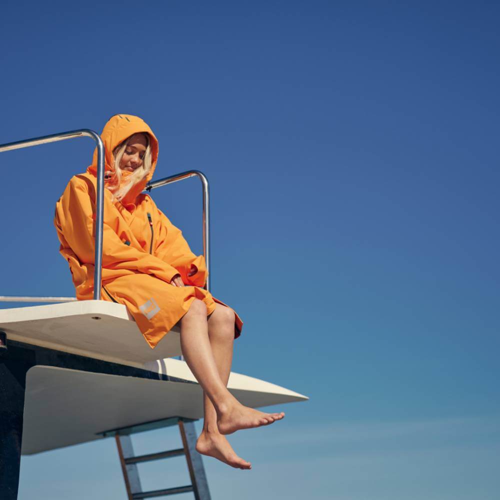 Woman sitting on the fly deck of a boat wearing a Red Alter Evo long sleeve waterproof changing robe in Bitter Orange.