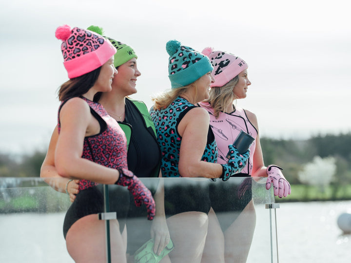 Four women wearing colorful beanies and swimming gear by a lake.