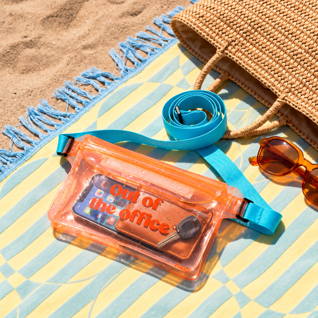 A clear orange waterproof bum bag with a bright blue strap, styled on a yellow and blue beach towel with sunglasses, a woven bag, and sand in the background.