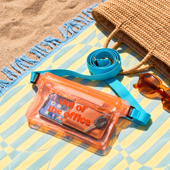 A clear orange waterproof bum bag with a bright blue strap, styled on a yellow and blue beach towel with sunglasses, a woven bag, and sand in the background.