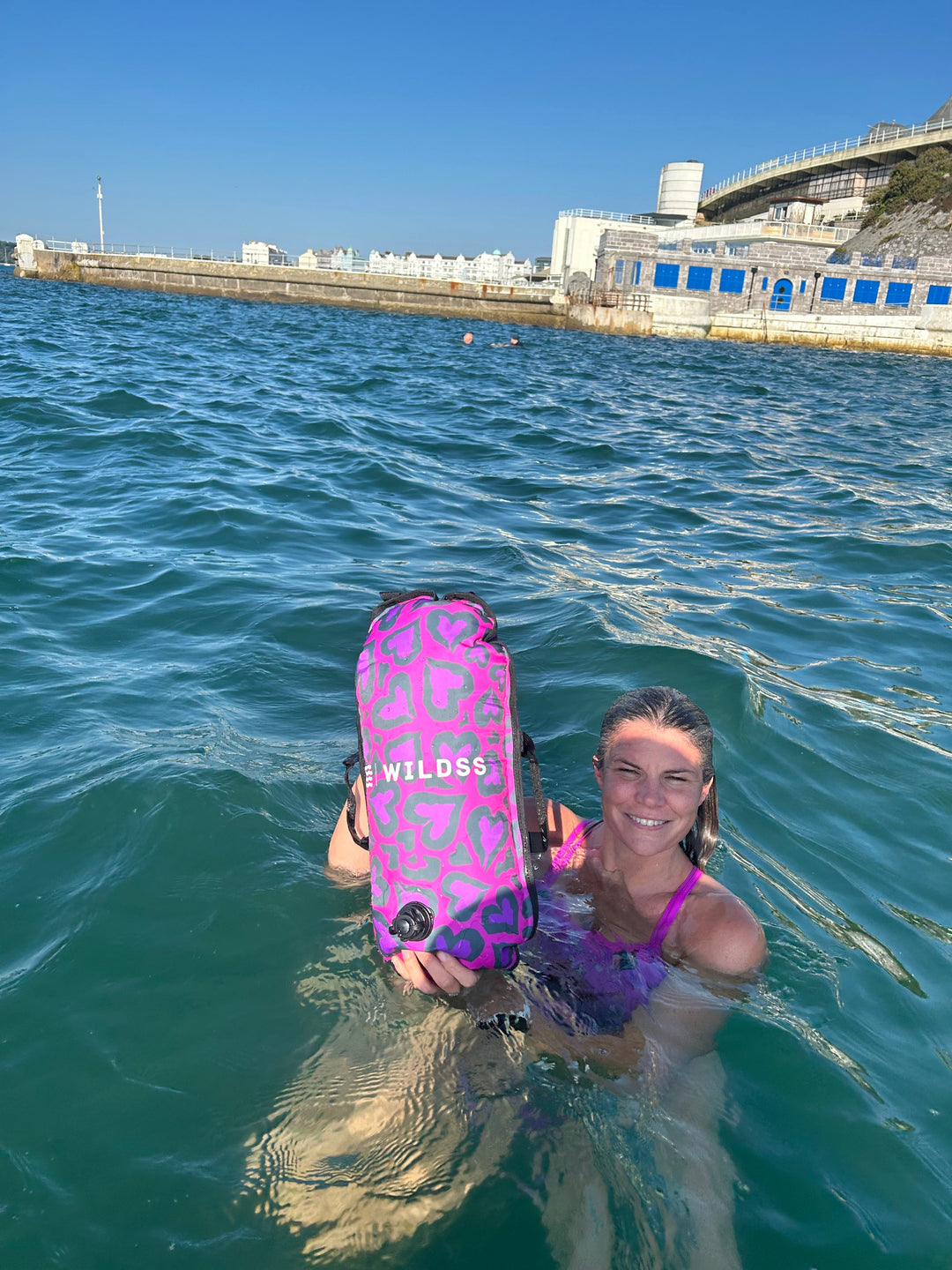 Swimmer in open water holding the Wild Swimming Socks 'Cold Cold Heart' print tow float with built-in thermometer, designed for open water safety.