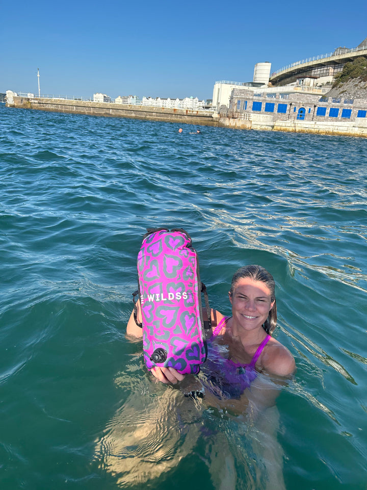 Swimmer in open water holding the Wild Swimming Socks 'Cold Cold Heart' print tow float with built-in thermometer, designed for open water safety.