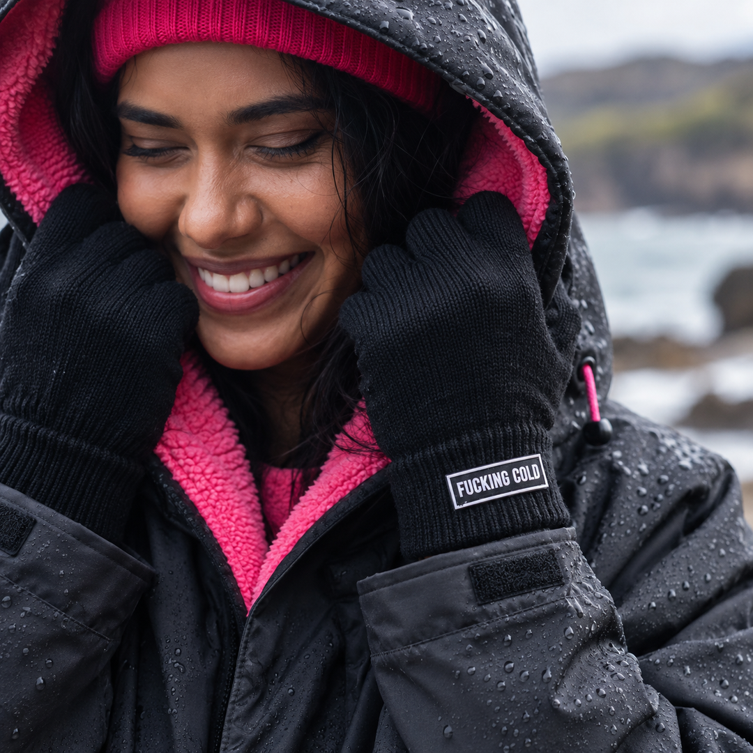 A woman wearing a black changing robe with a bright pink fleece lining and black gloves. The gloves have a small  rectangular patch on the cuff that reads 'FUCKING COLD' in white text.
