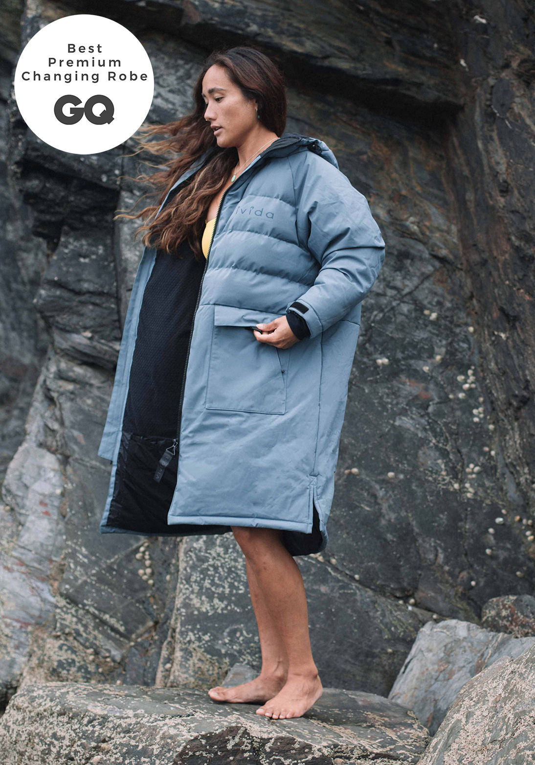 Woman wearing a Vivida Lifestyle All Weather Puffer Changing Robe, Mineral Blue Dry Robe for swimming standing on a rock. Sticker: Best Premium Changing Robe - GQ magazine.