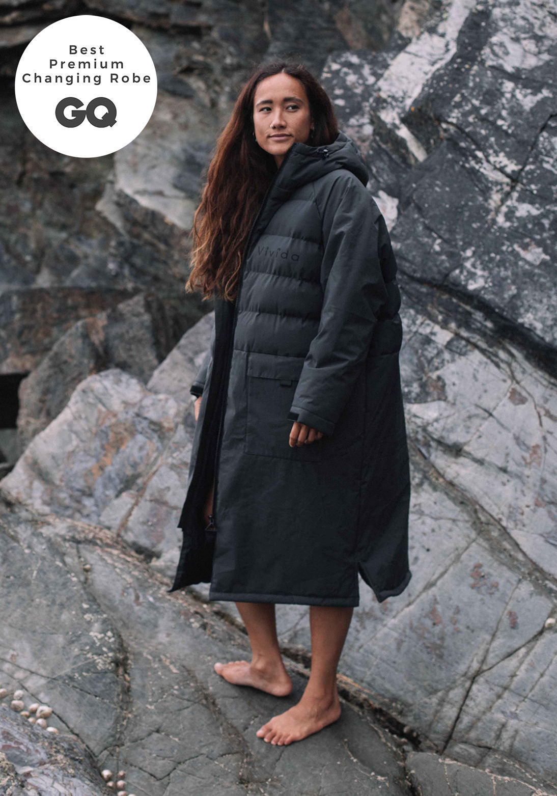 Woman wearing a Vivida Lifestyle All Weather Puffer Changing Robe, Fossil Grey Dry Robe for swimming standing on a rock. Sticker: Best Premium Changing Robe - GQ magazine.