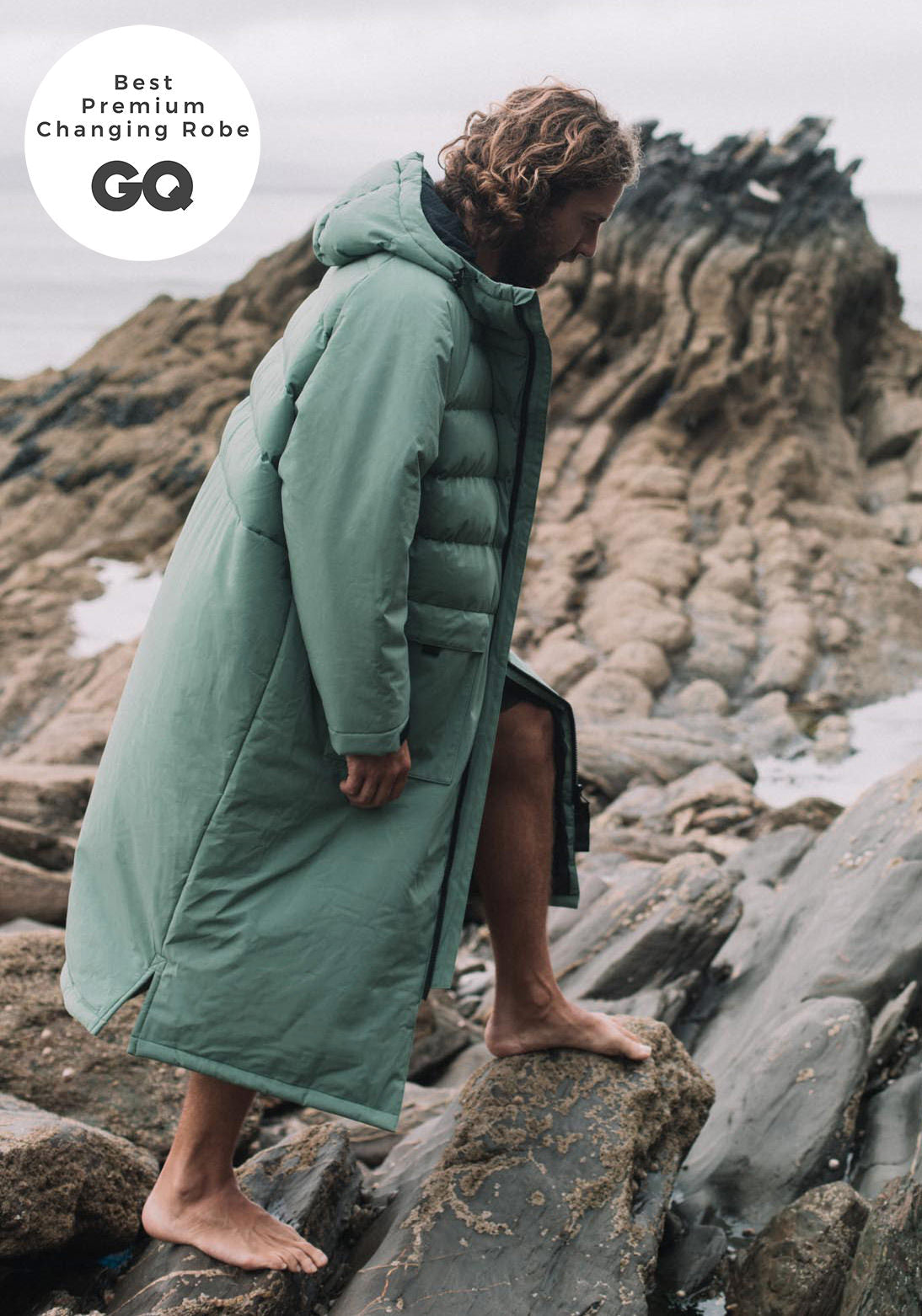 Woman wearing a Vivida Lifestyle All Weather Puffer Changing Robe, Aventurine Green Dry Robe for swimming standing on a rock. Sticker: Best Premium Changing Robe - GQ magazine.