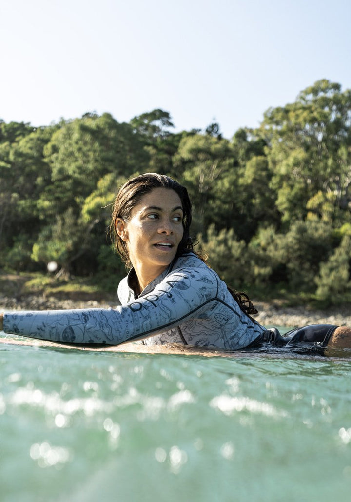 Lifestyle image of a woman lying on a surf board paddling into the sea wearing the Vivida Lindareversible neoprene swimsuit.